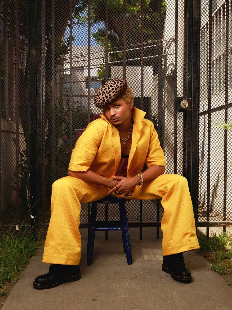 DESTIN CONRAD in yellow suit and leopard beret sits on stool in front of metal gate.