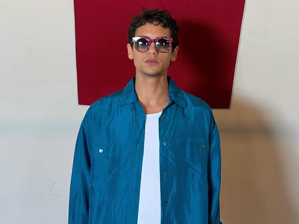 Dominic Fike wearing a teal shirt and sunglasses, standing against a red geometric background.