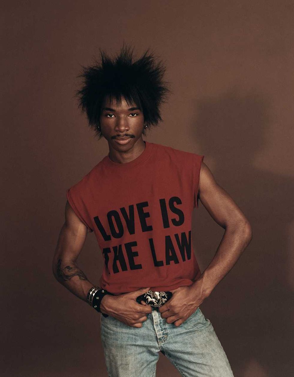 Love Spells with spiky hair in red "Love Is The Law" shirt and jeans against a brown background.