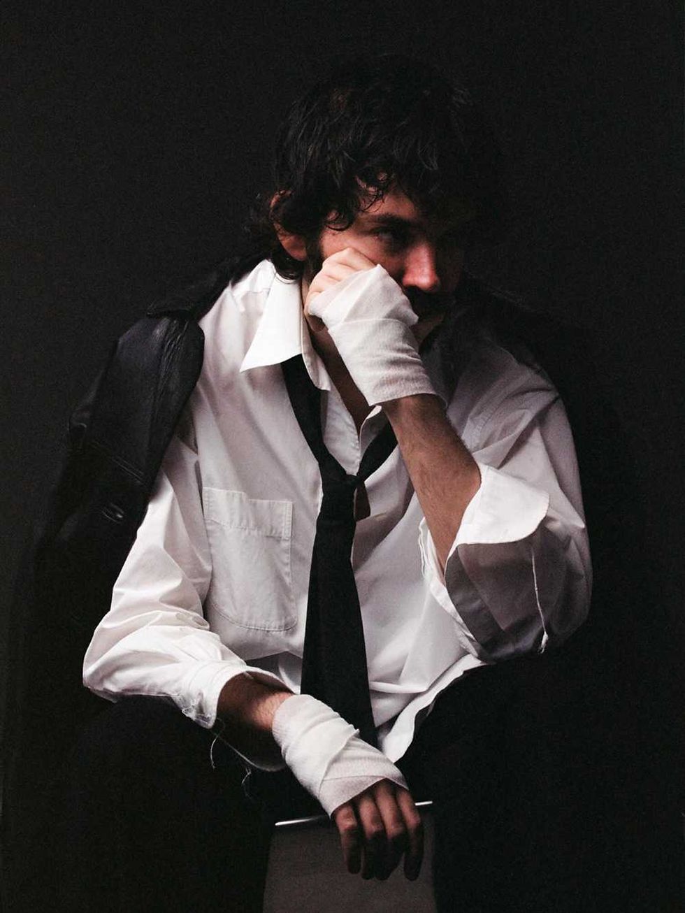 Madeon in white shirt and tie, wearing bandaged hands, sits contemplatively against a dark background.