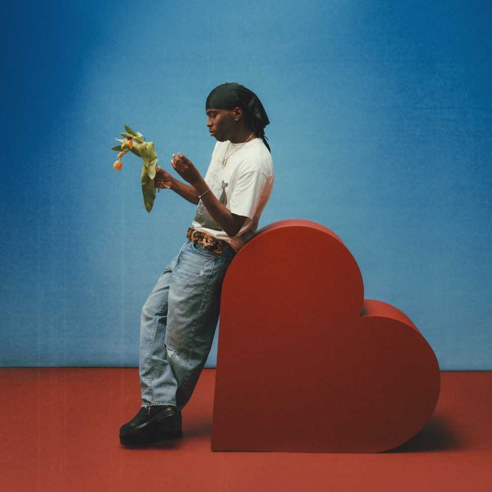 Master Peace leaning on red heart sculpture, holding tulips, against blue background.
