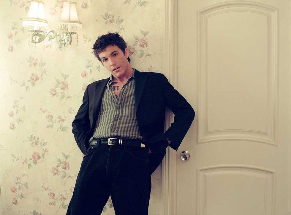 ROLE MODEL in a striped shirt and blazer leaning against a floral wallpapered wall.