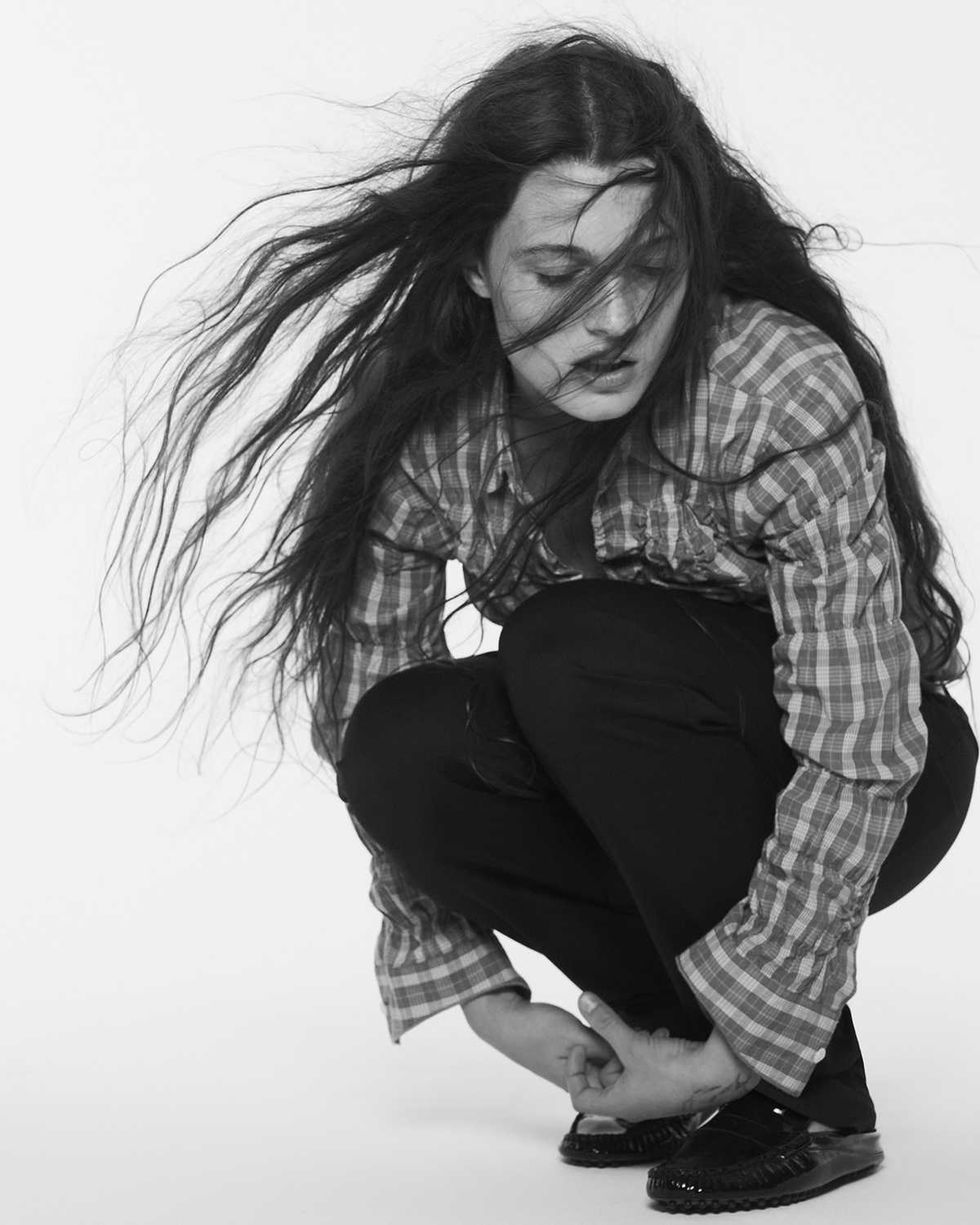 Sophia Stel in plaid shirt crouching, long hair blowing, eyes closed, black pants and shoes.