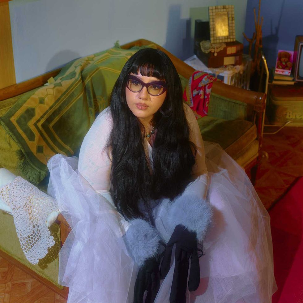 Sparklmami in glasses and furry gloves, seated on a vintage sofa with patterned cushions.